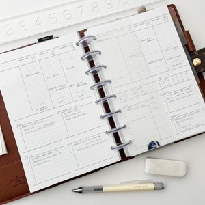 Peut inclure: Un planneur blanc ouvert sur une semaine, avec une couverture en cuir marron. Le planneur est ouvert sur une semaine avec des notes manuscrites et des listes de choses &agrave; faire. Le planneur est dot&eacute; d'un syst&egrave;me de reliure &agrave; disques en plastique transparent.