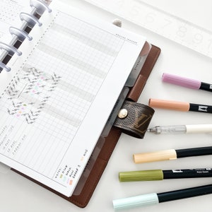 Peut inclure: Un planneur en cuir marron avec une page quadrill&eacute;e et un onglet intitul&eacute; "Routine Tracker". Le planneur est ouvert sur une page avec une section quadrill&eacute;e pour suivre les activit&eacute;s quotidiennes. La page comprend &eacute;galement une section pour les notes et une l&eacute;gende avec des points de diff&eacute;rentes couleurs repr&eacute;sentant diff&eacute;rentes activit&eacute;s. Il y a plusieurs marqueurs et stylos de couleurs sur une surface blanche &agrave; c&ocirc;t&eacute; du planneur.