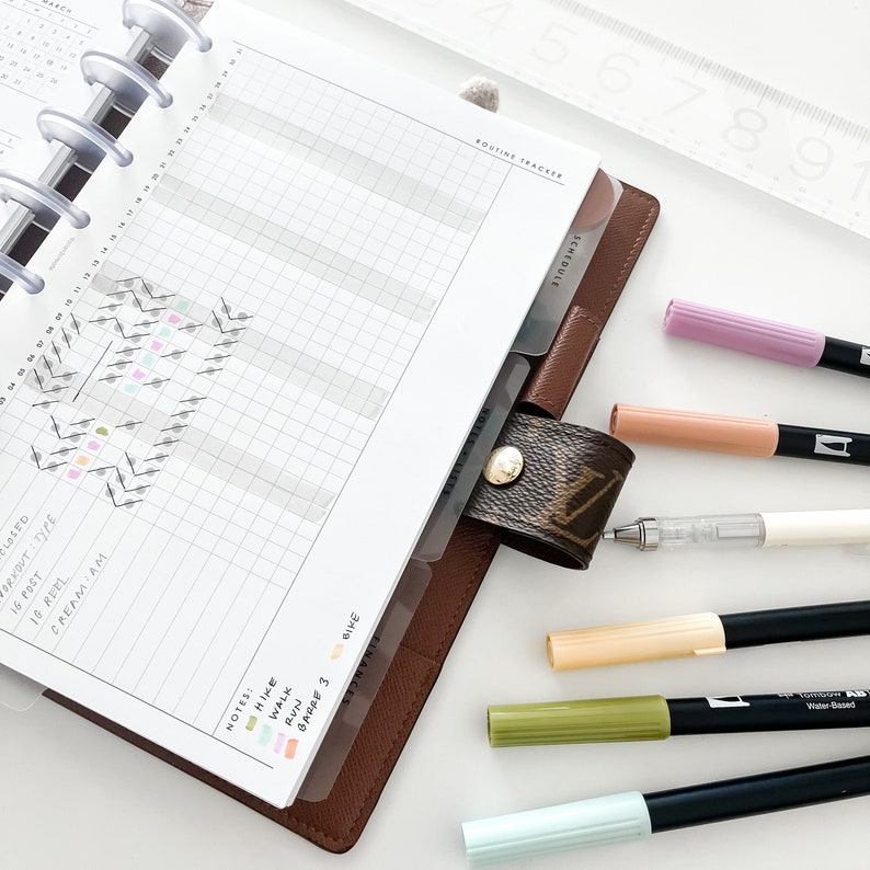 Peut inclure: Un planneur en cuir marron avec une page quadrill&eacute;e et un onglet intitul&eacute; "Routine Tracker". Le planneur est ouvert sur une page avec une section quadrill&eacute;e pour suivre les activit&eacute;s quotidiennes. La page comprend &eacute;galement une section pour les notes et une l&eacute;gende avec des points de diff&eacute;rentes couleurs repr&eacute;sentant diff&eacute;rentes activit&eacute;s. Il y a plusieurs marqueurs et stylos de couleurs sur une surface blanche &agrave; c&ocirc;t&eacute; du planneur.