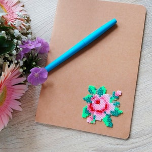 Op de afbeelding: Een bruin notitieboek met een blauwe pen erop. Een kleurrijke pixel art roos is op het notitieboek bevestigd. Roze gerbera's en paarse bloemen staan op de achtergrond.