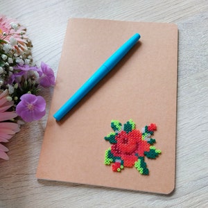 May include: A brown notebook with a blue pen resting on top. A red rose design made of small beads is affixed to the notebook. The notebook is on a light wood surface, with flowers in the background.