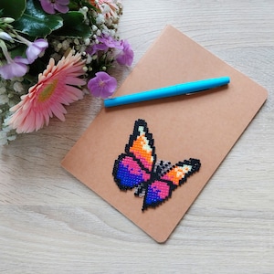 May include: A brown notebook with a colourful pixelated butterfly design. A blue pen rests on the notebook, and a bouquet of flowers is in the background. The butterfly features shades of pink, orange, and blue.
