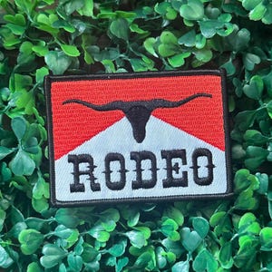 May include: Embroidered patch with a red and white background, featuring a black bull silhouette and the word "RODEO" in bold, black letters. The patch has a black border and is set against a backdrop of green foliage.