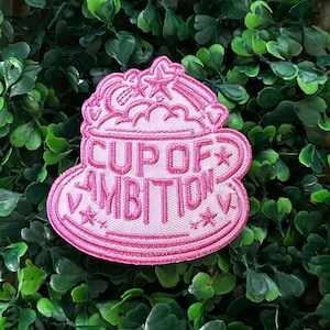 Peut inclure: Patch brodé rose à coller au fer avec un fond blanc représentant une tasse de café de dessin animé avec un nuage de vapeur et des étoiles. Le texte "Cup of Ambition" est brodé en rose sur la tasse.
