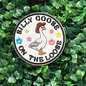 Op de afbeelding: Geborduurde patch met een cartoon gans met een bruine cowboyhoed. De patch heeft de tekst "SILLY GOOSE ON THE LOOSE" in zwarte letters. Het ontwerp bevat kleurrijke bloemen rond de gans.