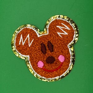Peut inclure: Un visage de souris en feutre marron avec un contour de paillettes dorées. La souris a un sourire rouge, des joues roses et des yeux noirs.