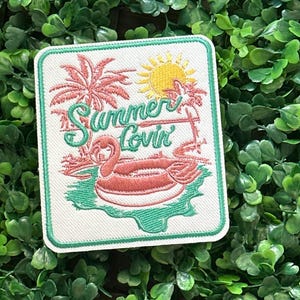 May include: Embroidered patch with a summer theme. The patch features a pink flamingo in a pool float, palm trees, and a yellow sun. The words "Summer Lovin'" are written in green script. The patch has a white background with a green border.