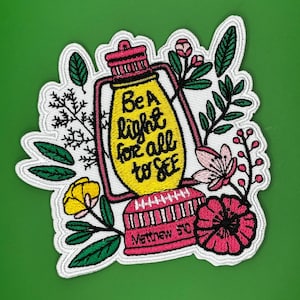 Puede incluir: Un parche bordado para planchar con una linterna amarilla y flores rosas. La linterna tiene el texto "Sé una luz para todos" y "Mateo 5:16".