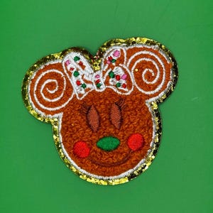 Peut inclure: Un écusson en forme de tête de souris façon pain d'épices, avec un visage marron et des détails brodés. L'écusson a un nœud blanc avec des accents colorés, des joues rouges et un nez vert. Les bords sont soulignés de paillettes dorées.