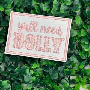 May include: A white patch with pink stitching and the text "y'all need Dolly" in pink lettering.