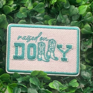 May include: A rectangular patch with a white background and a teal border. The patch features the text "raised on DOLLY" in teal script and block letters, with a pair of teal cowboy boots in the center. The patch is surrounded by green foliage.