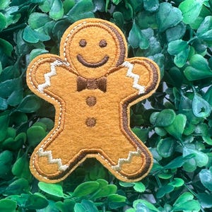 以下が含まれることがあります： 笑顔、蝶ネクタイ、装飾的なステッチが施された、陽気なジンジャーブレッドマンのパッチ。パッチは温かみのある茶色で、白と茶色のアクセントがあります。背景は緑の葉のベッドです。