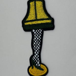 May include: A yellow lampshade with a black base and a black and white fishnet stocking leg with a black high heel shoe. The lampshade has a green trim.