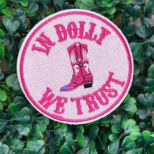 以下が含まれることがあります： ピンクの刺繍パッチに、ピンクの カウボーイブーツと「In Dolly We Trust」の文字が刺繍されています。