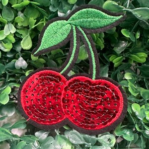 May include: Embroidered cherry patch with red sequin detailing, black outline, and green leaves. This patch is a fun accessory for clothing or bags, adding a pop of colour and a touch of whimsy.