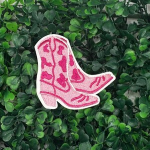 May include: Pink cowboy boot embroidered patch with white stitching. The boots have a pink and white pattern.