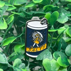 Puede incluir: Un pin de esmalte azul marino con forma de lata de bebida, con una niña bajo un paraguas bajo la lluvia. El pin tiene una parte superior blanca y la palabra "SALTY" en amarillo. El pin está rodeado de hojas verdes.