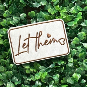 May include: A rectangular white patch with a brown border and the embroidered phrase "Let them" in brown script, with a small heart above the "m". The patch is set against a backdrop of green foliage.