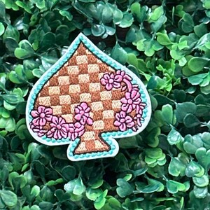May include: A decorative patch in the shape of a spade, featuring a brown checkered pattern and pink floral accents. The spade is outlined in a light blue color, with a white border. The patch is set against a backdrop of green foliage.