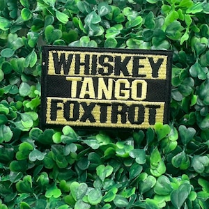 May include: A black and gold embroidered patch with the words "WHISKEY TANGO FOXTROT" in bold, yellow letters. The patch is rectangular and has a black border. The patch is set against a background of green foliage.