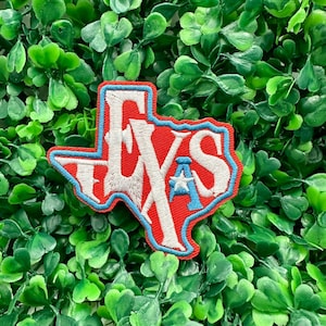 Puede incluir: Parche bordado rojo, blanco y azul con la forma del estado de Texas. El parche presenta la palabra "TEXAS" en blanco, con un contorno azul y una estrella azul. El parche está sobre un fondo de hojas verdes.