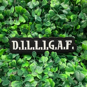 May include: A black rectangular patch with white text that reads "D.I.L.L.I.G.A.F." The patch is set against a background of vibrant green foliage. The patch is likely an accessory for clothing or bags.