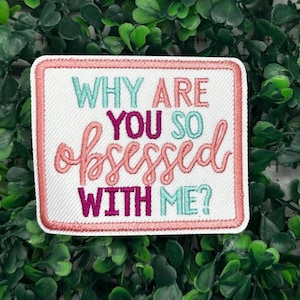 Pode incluir: Um remendo branco bordado com letras rosa, roxas e azuis que diz "Why are you so obsessed with me?"
