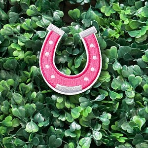 May include: A pink horseshoe patch with white polka dots and silver accents. The patch is set against a backdrop of green foliage. The horseshoe is a symbol of good luck and is often used in crafts and apparel.