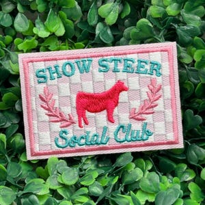 Puede incluir: Parche rectangular con las palabras "SHOW STEER Social Club" bordadas en color turquesa. Un toro rojo está centrado en un fondo a cuadros rosa y blanco, enmarcado por hojas rosas y un borde rosa.