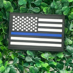 Può includere: Una toppa in bianco e nero con il design della bandiera americana con una striscia blu. La toppa ha stelle e strisce bianche, con una striscia blu al centro. La toppa è rettangolare e posta su uno sfondo di fogliame verde.