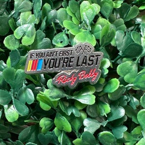 May include: A silver pin with the text "If you ain't first you're last - Ricky Bobby" in red and black lettering. The pin also features a checkered flag graphic.