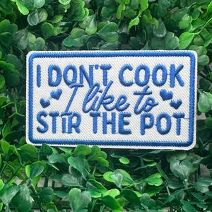 Puede incluir: Parche rectangular con fondo blanco y borde azul. Presenta el texto bordado en azul: "I DON'T COOK I like to STIR THE POT", con pequeños acentos de corazón azul. El parche está sobre un fondo de follaje verde.