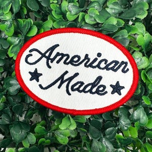 Puede incluir: Parche ovalado blanco con costuras rojas alrededor del borde. El parche tiene el texto "American Made" en letras cursivas negras con una estrella a cada lado de la palabra "Made".