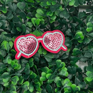 May include: Pink heart-shaped sunglasses with silver sequins. The sunglasses are on a green leafy background.