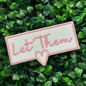Puede incluir: Un parche rectangular blanco con texto bordado en rosa que dice "Let Them". Una forma de corazón rosa está debajo de la palabra "Them". El parche tiene un borde rosa y está sobre un fondo de hojas verdes.