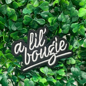 Op de afbeelding: Zwart geborduurde patch met de woorden "lil bougie" in witte letters. De patch heeft een zwarte rand en is geplaatst tegen een achtergrond van groen gebladerte.