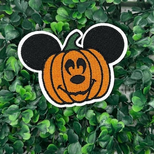 May include: An orange pumpkin with a Mickey Mouse face, black ears, and a green stem. The pumpkin is embroidered on a white background.