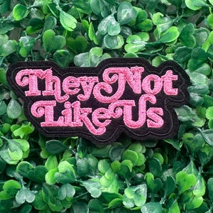 May include: A black patch with the embroidered text "They Not Like Us" in pink, with a decorative font. The patch is set against a background of green foliage.