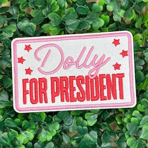 May include: Embroidered patch with the words "Dolly FOR PRESIDENT" in pink and red thread. The patch is rectangular with a white background and a pink border. Red star accents are placed around the text. The patch is set against a backdrop of green foliage.