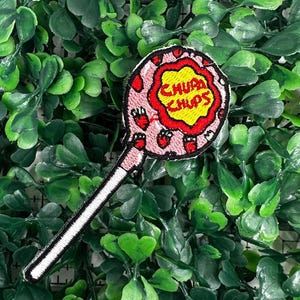 May include: A pink and white embroidered iron-on patch of a lollipop with the text "Chupa Chups" in yellow.