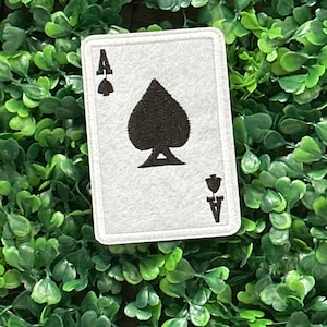 Puede incluir: Un parche blanco con el diseño de la carta del As de Picas. La carta tiene un símbolo de pica negro en el centro, con la letra "A" y un pequeño símbolo de pica en cada esquina. El parche está rodeado por un borde blanco.
