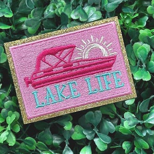 May include: Pink embroidered patch with a gold glitter border. The patch features a pink boat with a white sun and the text "LAKE LIFE" in turquoise.