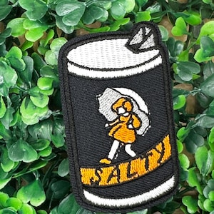 Puede incluir: Parche bordado negro de una lata con una niña sosteniendo un paraguas y la palabra "Salty" en él.