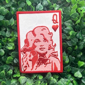 May include: A rectangular patch with a red border features a red and white image of a woman's face, resembling a playing card. The top right corner displays a red heart and the letter "Q". The patch is set against a backdrop of green foliage.