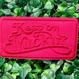 以下が含まれることがあります： スタイリッシュな筆記体フォントで「Keep on Truckin'」というフレーズが刺繍された、長方形の赤いパッチ。パッチは質感があり、ステッチされた縁取りがあります。背景は緑の葉のベッドです。