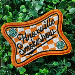 Könnte beinhalten: Orangefarbener und weißer karierter Patch mit dem Text "Knoxville Smokershow" in schwarzer Kursivschrift. Der Patch hat einen dekorativen orangefarbenen Rand und zwei blaue Blumenmuster.