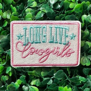 May include: Embroidered patch with the text "LONG LIVE Cowgirls" in turquoise and pink. The patch has a light beige background with a pink border and two turquoise stars. The patch is rectangular in shape.