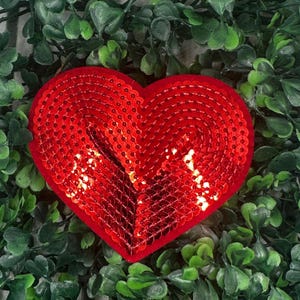 May include: A red heart-shaped sequin patch. The sequins are arranged in a pattern that creates a three-dimensional effect.