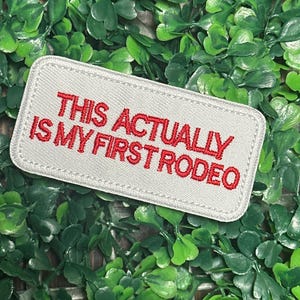 Könnte beinhalten: Ein rechteckiger, hellgrauer Aufnäher mit rotem, gesticktem Text: "THIS ACTUALLY IS MY FIRST RODEO." Der Aufnäher hat einen weißen, genähten Rand, vor einem Hintergrund aus grünem Laub.