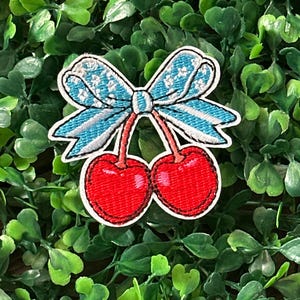 Op de afbeelding: Geborduurde patch met twee rode kersen met groene stelen, verbonden met een blauwe strik met witte sterdetails. De patch heeft een witte rand en is geplaatst tegen een achtergrond van groen gebladerte.